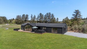 A modern black house with a large glass front, a deck, and a patio with outdoor furniture. - Beautiful home in Hjørring with sauna (Hjørring)