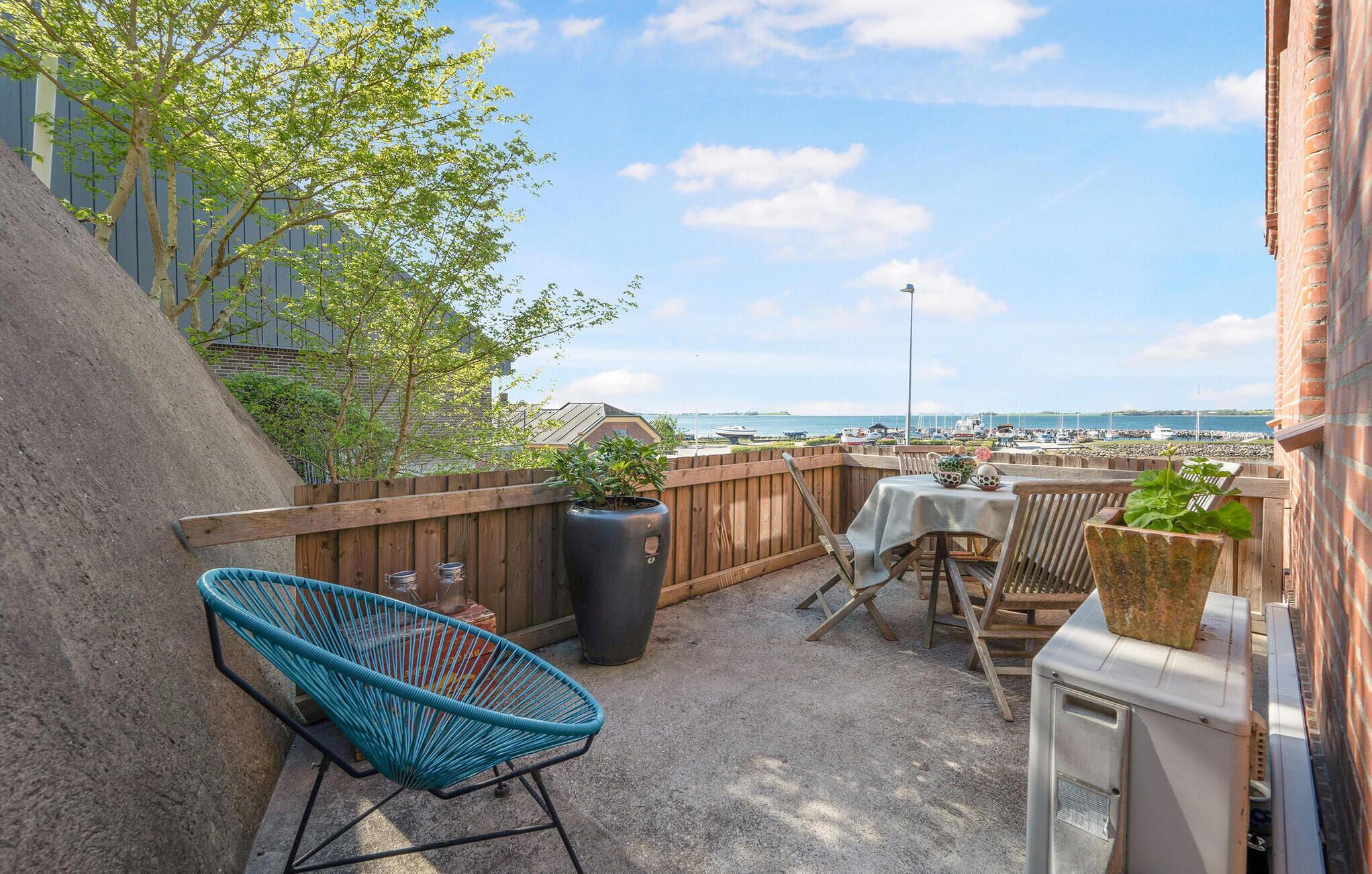 A patio with a view of the sea, a wooden fence, a blue chair, a table with chairs, and a potted plant.