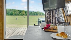 A table with a croissant, a plate, a cup, and a tray with grapes and bread, with a TV mounted on the wall and a view of a grassy field through large windows. - 1 bedroom beautiful home in Kolding (Kolding)