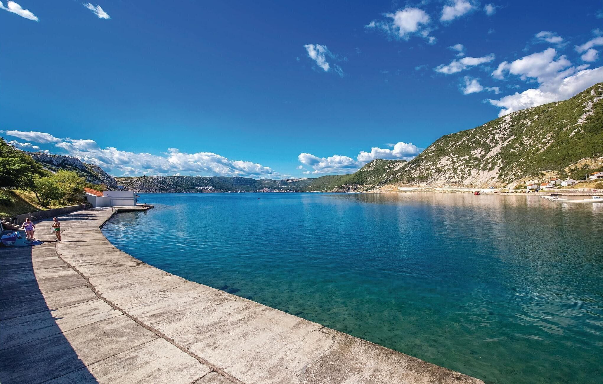 Una escena frente al agua con un muelle de hormigón, agua azul cristalina y un paisaje montañoso al fondo.