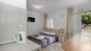 Habitación de hotel compacta con cama, mesita de noche, una pequeña mesa, hervidor, ventilador y televisor.