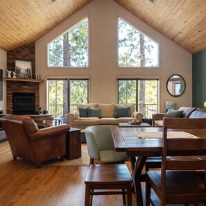 A spacious living room with a high ceiling, large windows, a fireplace, and a wooden dining table.