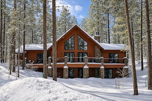 A log cabin with a stone foundation, large windows, and a balcony, surrounded by a snowy forest.