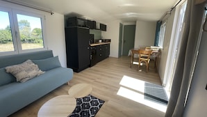 Uma sala de estar moderna com um sofá azul, uma mesa de jantar com cadeiras, uma cozinha com armários pretos e micro-ondas, e uma janela grande que oferece iluminação natural.