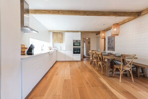 A modern kitchen with white cabinetry, wooden beams, and a dining area with a wooden table and chairs. - The Cottage at The Pines, Bull Bay. (Anglesey)
