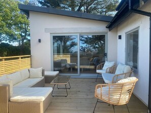 A modern outdoor patio with a wooden deck, wicker furniture, and a sliding glass door leading to an adjacent room.