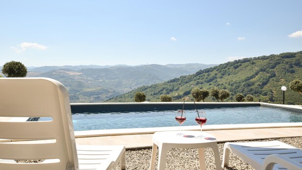Una piscina al aire libre con dos sillas reclinables blancas y una mesa, con vistas a un paisaje montañoso.