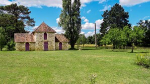 Una pequeña iglesia de piedra con techo de tejas y puertas moradas, rodeada por un campo cubierto de césped y árboles.