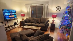 Un salon avec un canapé, un pouf, une table basse, un téléviseur et un sapin de Noël.
