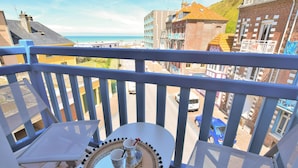 Un balcon avec une table dressée pour deux personnes, donnant sur une ville côtière avec ses bâtiments et un ciel dégagé.