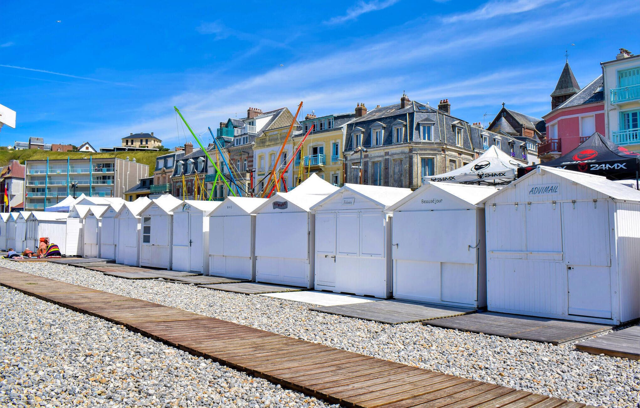 Eine Reihe weißer Strandhütten mit einem hölzernen Steg, der zu ihnen führt, vor einer Kulisse aus bunten Gebäuden und einem klaren, blauen Himmel.