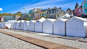 Une rangée de cabanes de plage blanches, avec une promenade en bois qui y mène, se dressant sur fond de bâtiments colorés et d’un ciel bleu azur.