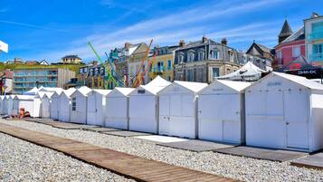 Eine Reihe weißer Strandhütten mit einem hölzernen Steg, der zu ihnen führt, vor einer Kulisse aus bunten Gebäuden und einem klaren, blauen Himmel.