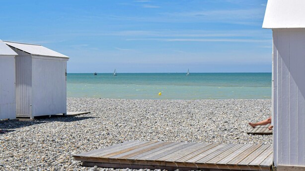 Une plage avec une terrasse en bois, des galets et un ciel bleu azur.