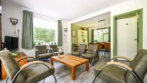 A living room with a wooden coffee table, green upholstered chairs, a television, and a dining area with a table and chairs.