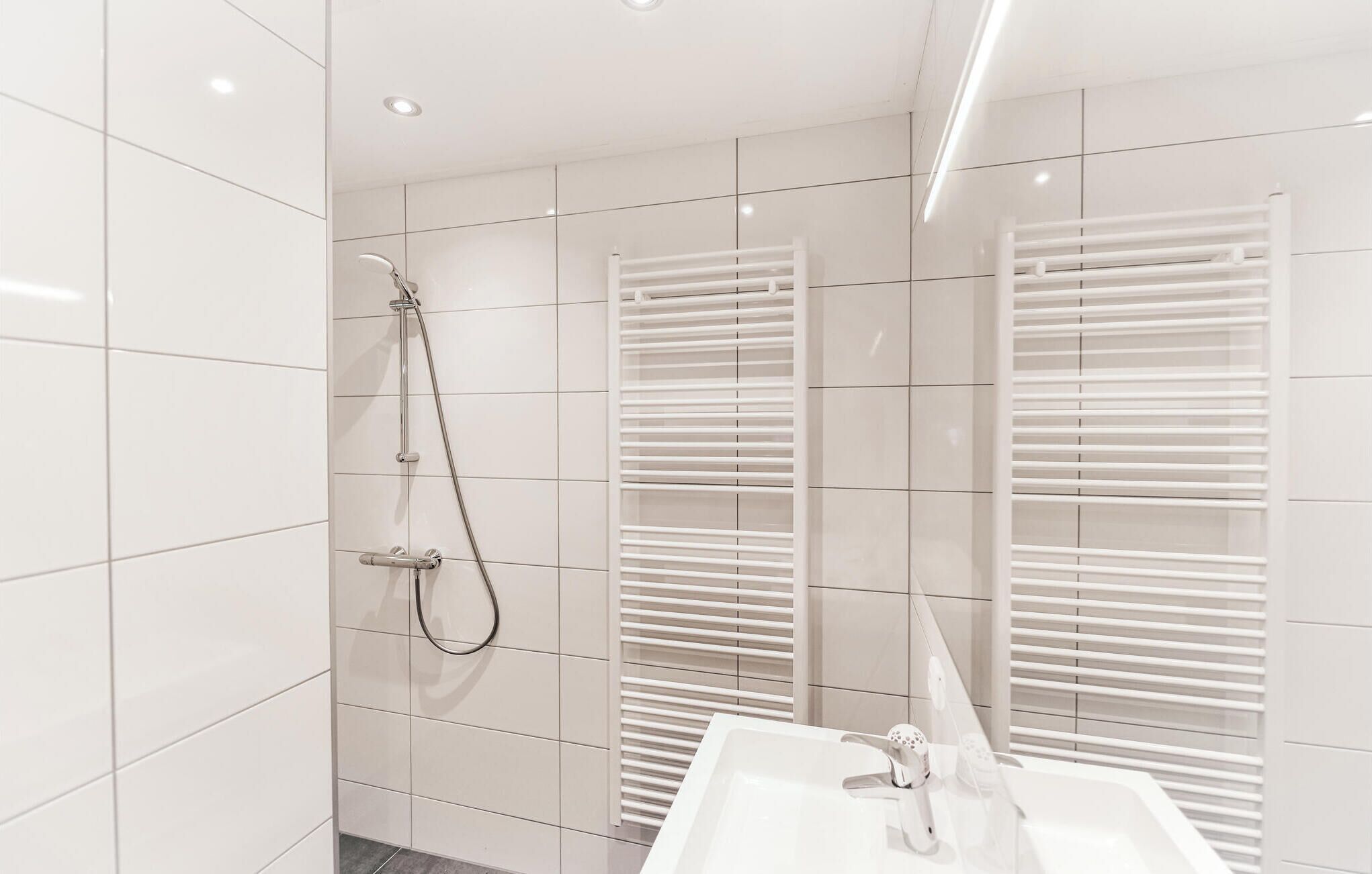A modern bathroom with white tiles, a shower with a handheld showerhead, and a white sink with a mirror.