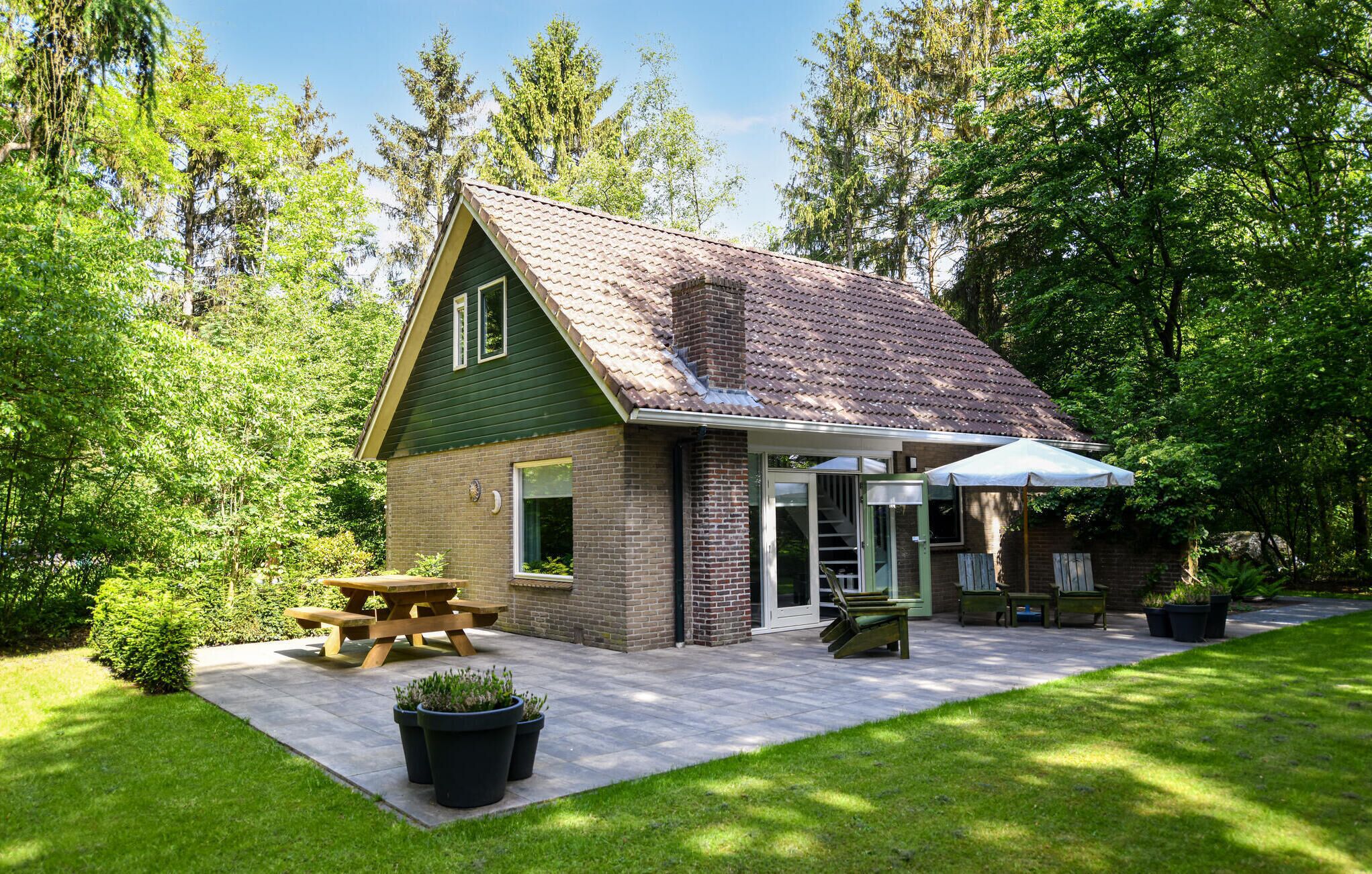 Eine kleine, eingeschossige Holzhütte mit grünem Dach und Backsteinwänden, eine Terrasse mit Picknicktisch und Stühlen sowie ein überdachter Sitzbereich im Freien.