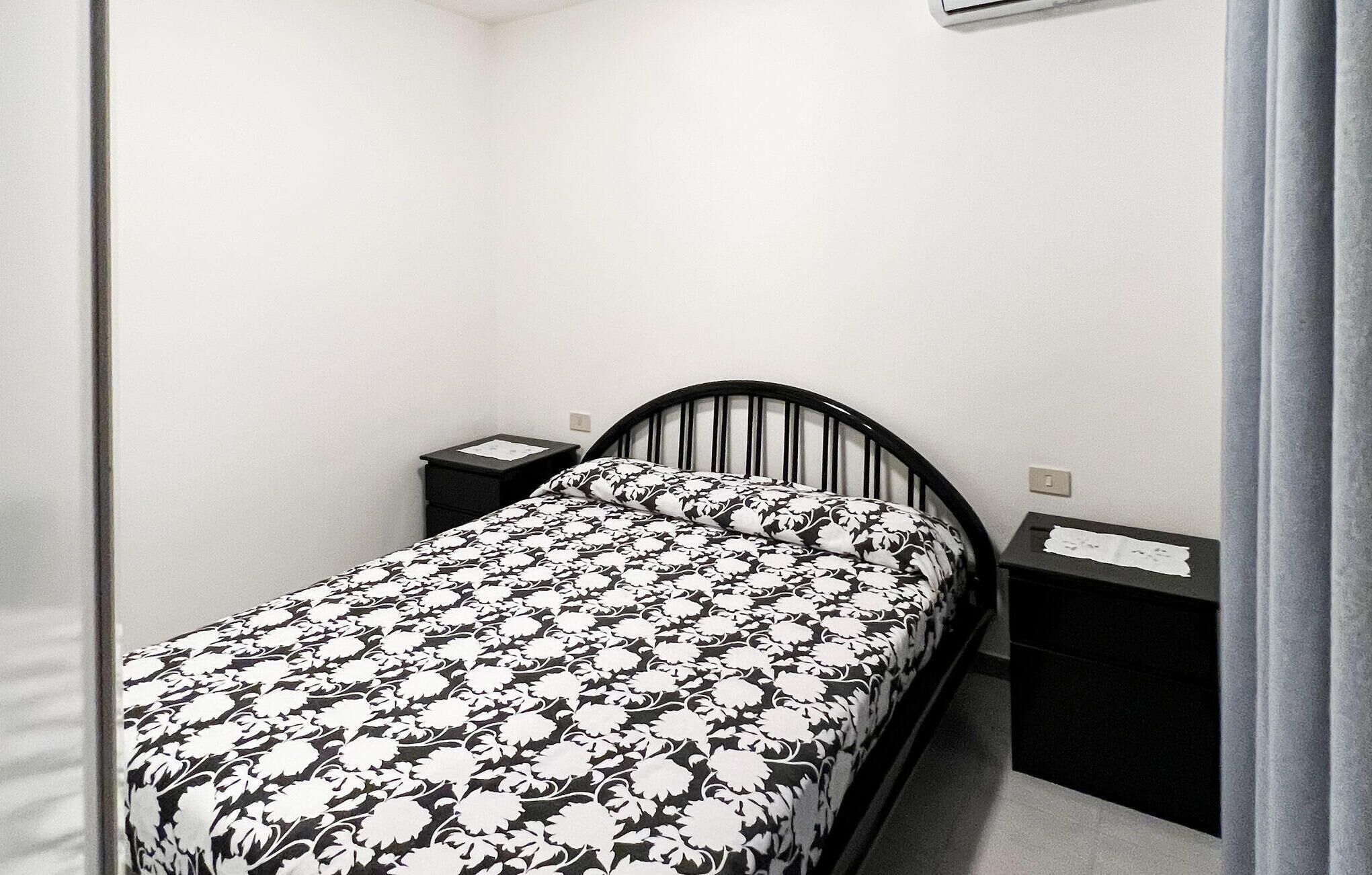 Una habitación pequeña con una cama individual y colcha de flores en blanco y negro, dos mesitas de noche con papeles, y una pared blanca lisa.