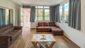 Ein Wohnzimmer mit braunem Ledersofa, einem Holztisch und großen Fenstern mit grauen Vorhängen.
