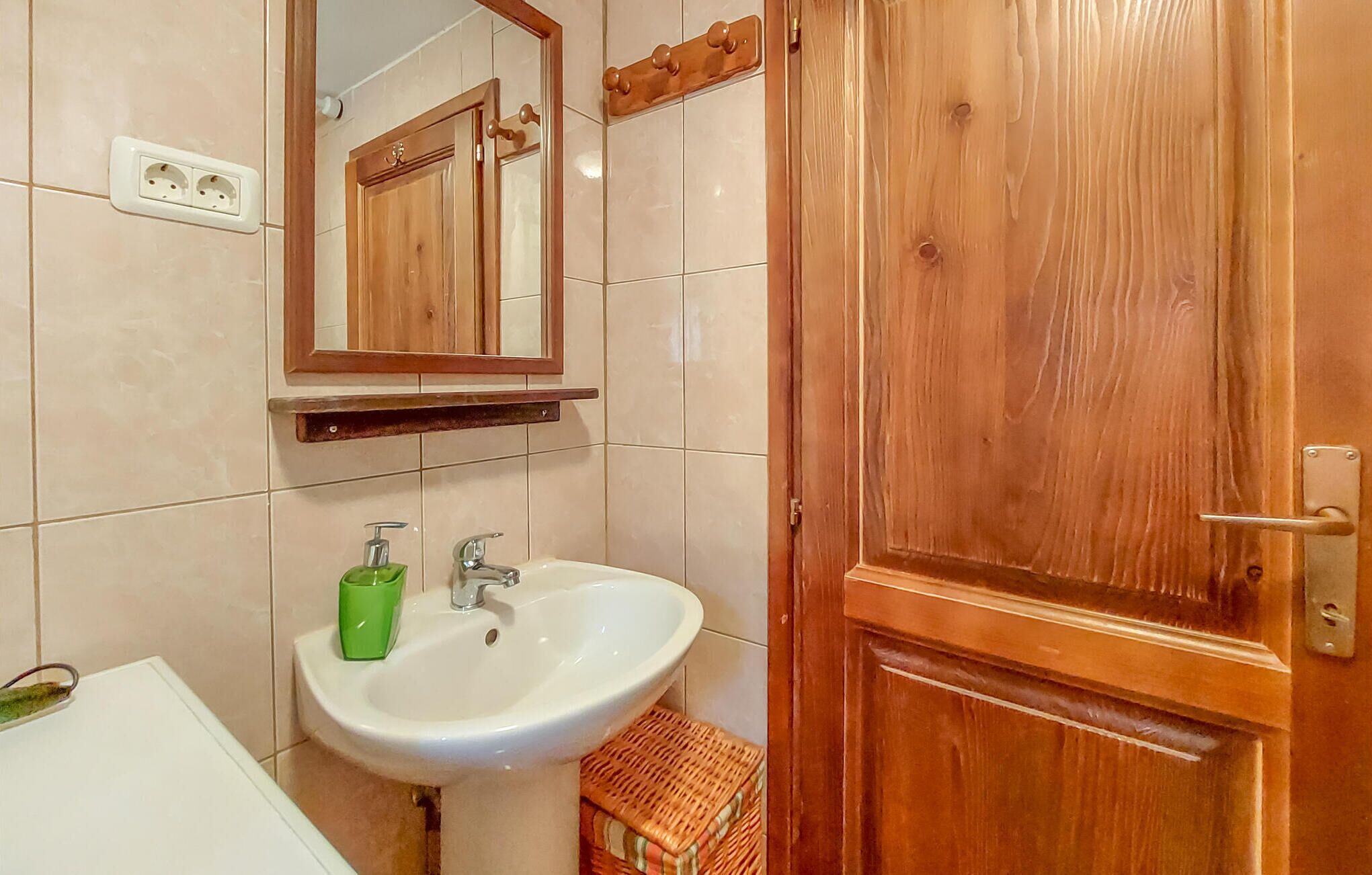A bathroom with a wooden door, a white sink with a green soap dispenser, and a mirror above the sink.