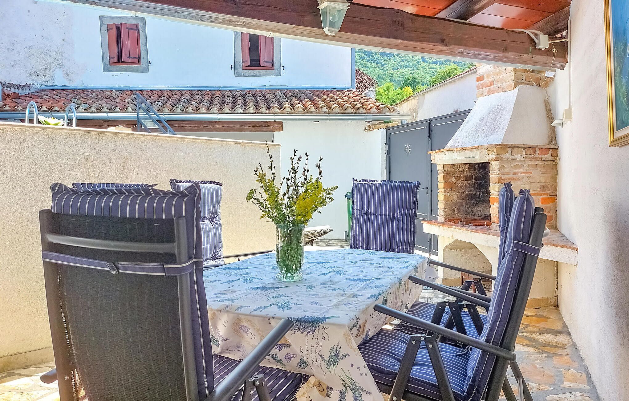 An outdoor patio with a table and chairs, a small brick oven, and a view of a building with red shutters.