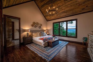 A bedroom with a large bed, a wooden ceiling, a view of the outdoors, and a nightstand with a lamp. - Gem of Blue Ridge | Pool + Views + Hot Tub (Blue Ridge)