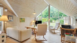 L’intérieur chaleureux d’un chalet avec un plafond en bois, de grandes fenêtres, un téléviseur et un petit coin repas.