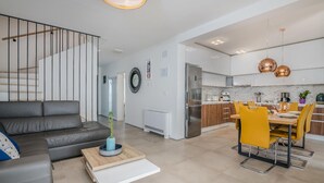 A modern living room with a grey sofa, a dining area with yellow chairs, and a kitchen with white cabinets and copper pendant lights.