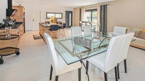 A modern dining area with a glass table and white chairs, a kitchen in the background, and a sofa in the living area. - Beautiful home in Robion (Robion)