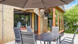 Eine Terrasse im Freien mit Tisch und Stühlen, einem großen Sonnenschirm und einer Glastür, die zu einem Garten führt.