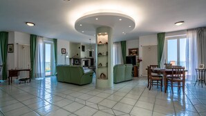 Amplia sala de estar con un pilar central, sofás verdes, una mesa de comedor con sillas y una chimenea.