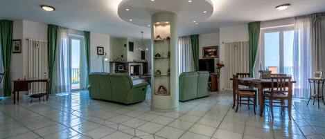A spacious living area with a central column, green sofas, a dining table with chairs, and a fireplace.