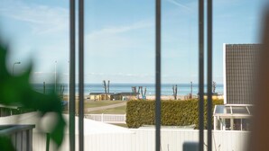 A view of a beach from a window with a white fence and a hedge in the foreground.