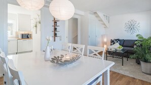 A modern kitchen with white cabinets, a microwave, and a dining table with chairs. There is a staircase leading to another level, a living area with a sofa and a coffee table, and a decorative wall piece.