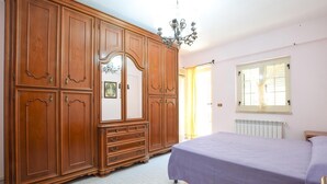 A bedroom with a large wooden wardrobe, a bed with a purple bedspread, a chandelier, and a window with blinds. - 1 bedroom nice apartment in Marina di Caronia (Marina di Caronia)