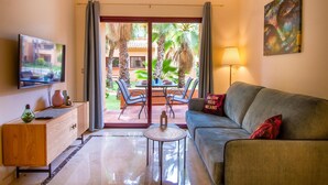 A living room with a sofa, a coffee table, a TV, and a view of an outdoor patio with chairs and palm trees.