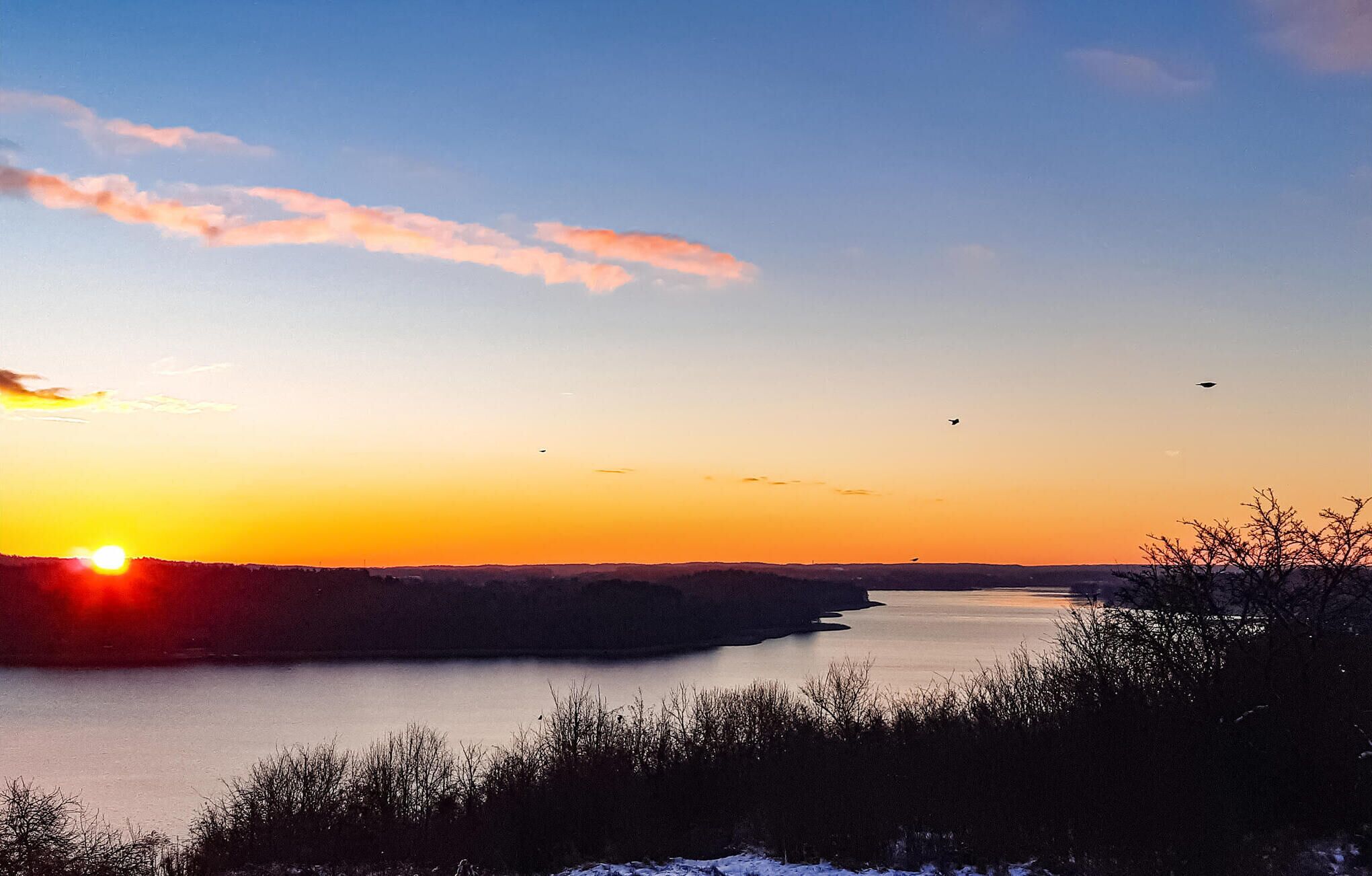A sunset over a calm lake with a distant shoreline and sparse vegetation.