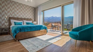 A bedroom with a large bed, a balcony with a view of mountains, and a teal armchair.