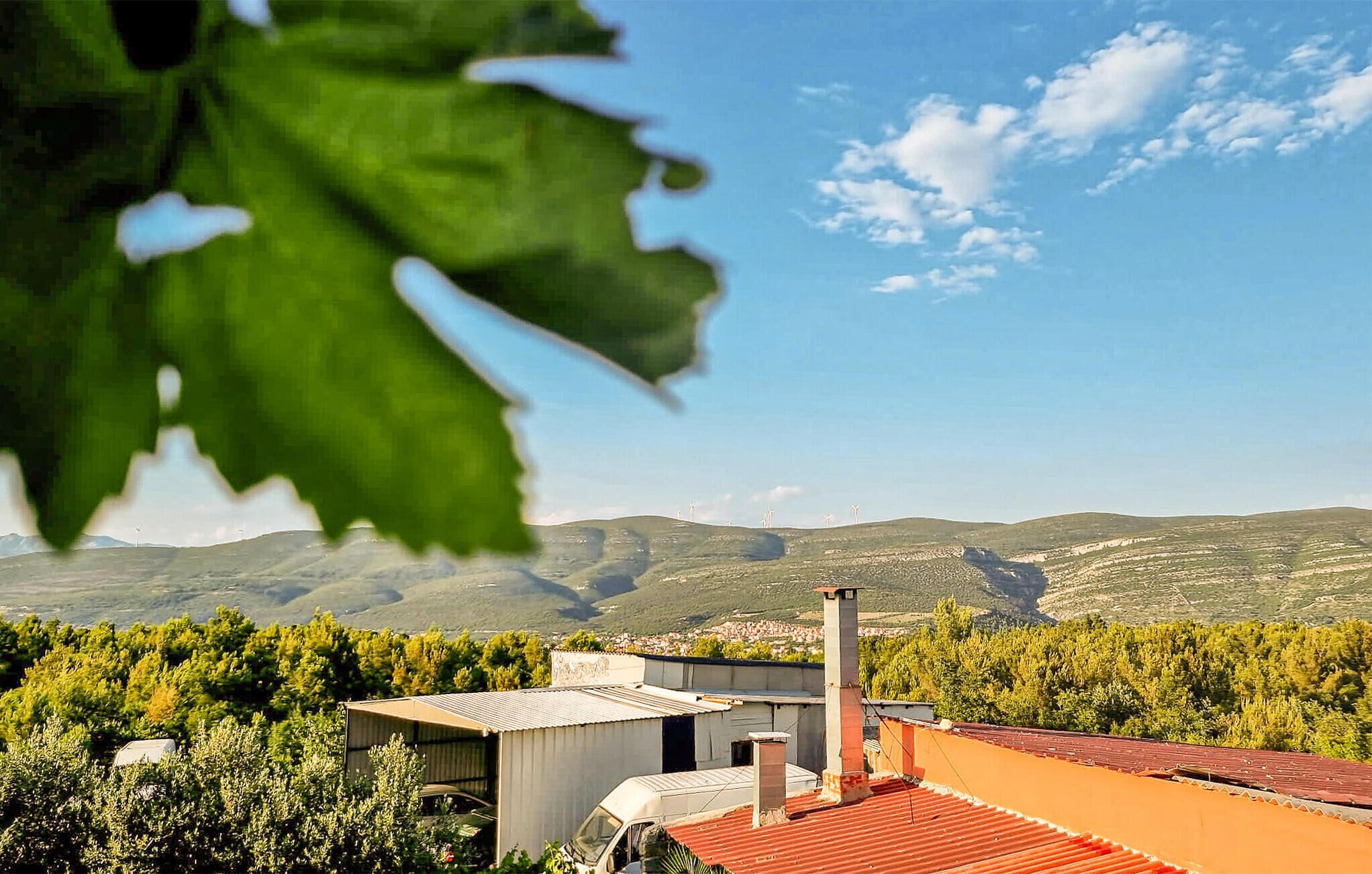 A rural landscape with a small building, possibly a house or a barn, surrounded by greenery and hills in the background.