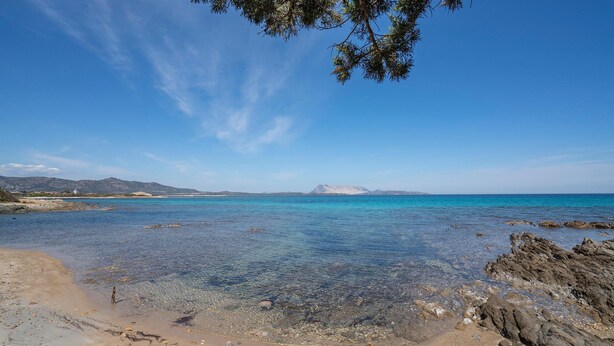 Een strand met helder turquoise water, een rotsachtige kustlijn en verre bergen.