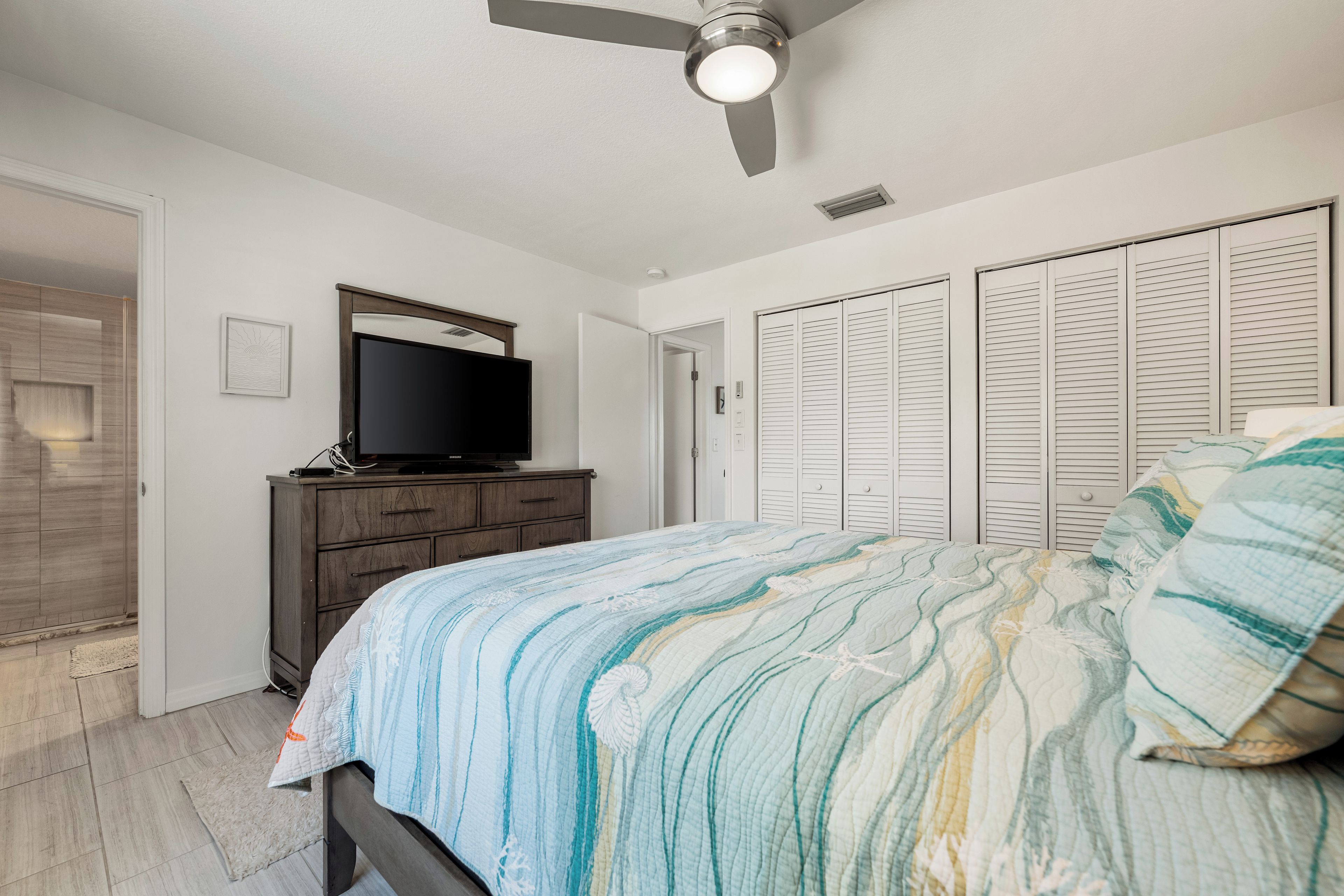 Une chambre à coucher avec un lit, un téléviseur posé sur une commode, un ventilateur de plafond et un placard avec des portes à persiennes.