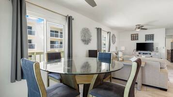Un salon moderne avec une table à manger en verre, des sièges confortables, un téléviseur à écran plat et un balcon offrant une vue sur un immeuble.