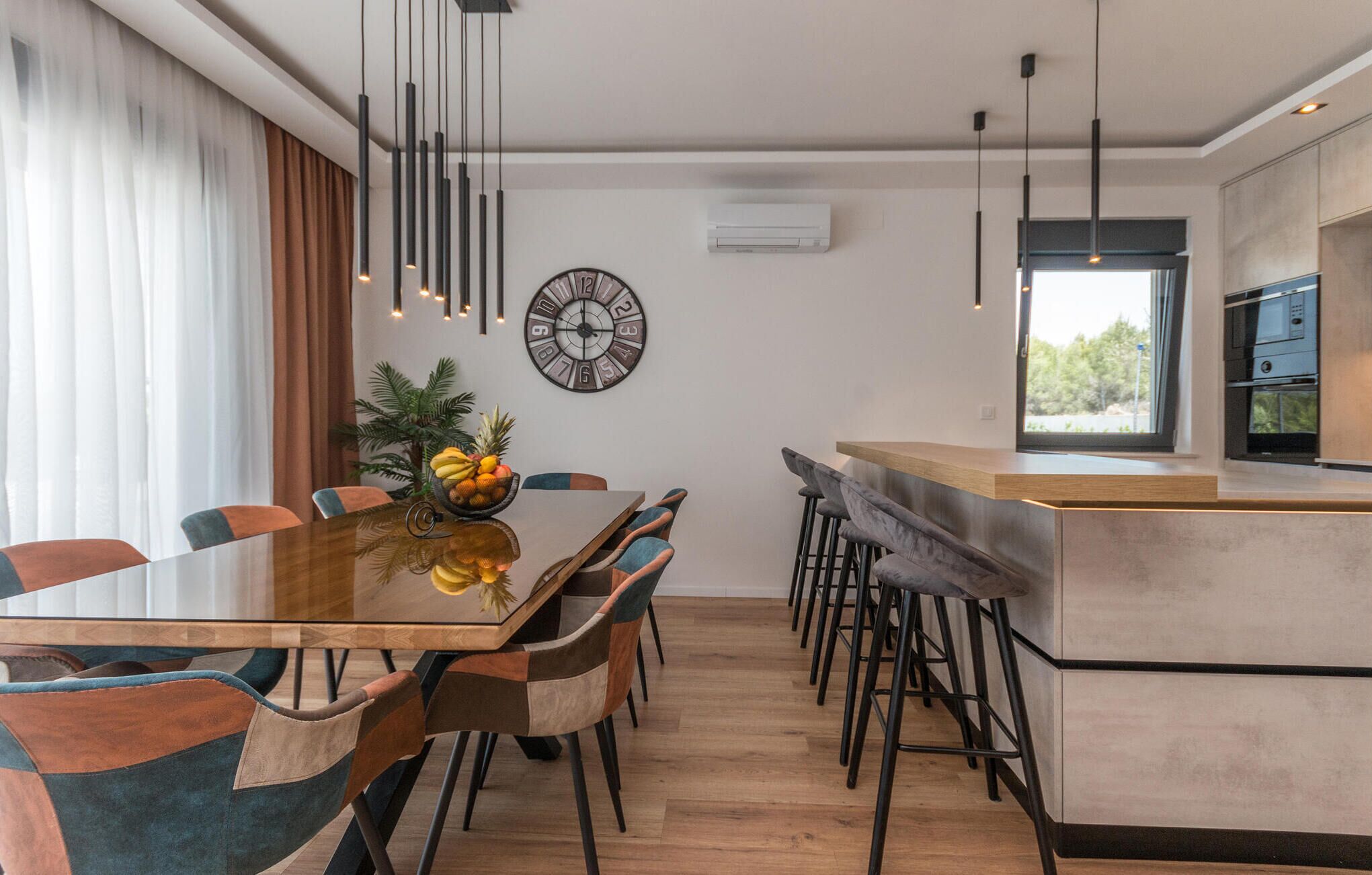 A modern kitchen with a central island, a dining area with a wooden table and chairs, and a large wall clock.