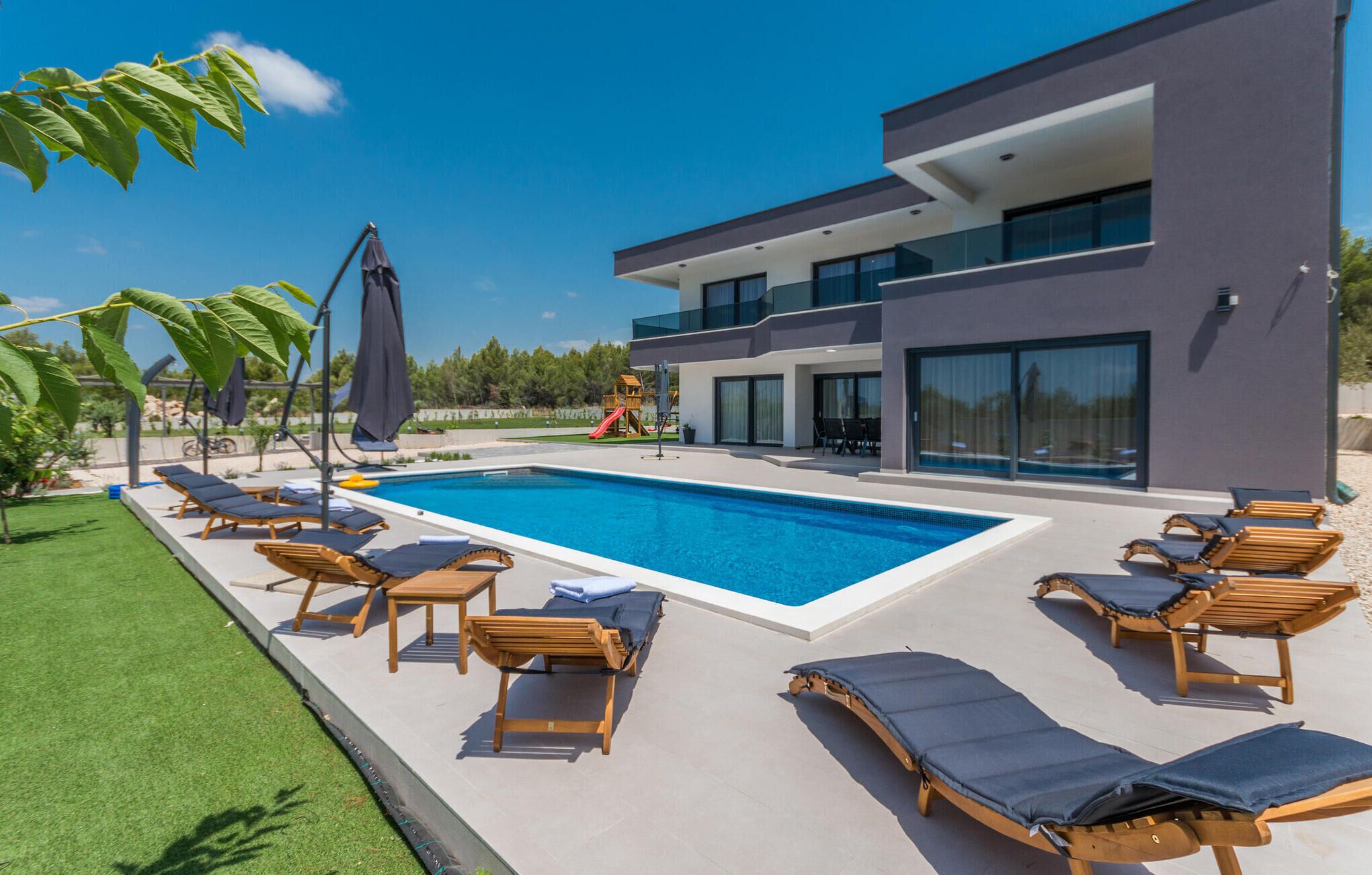 A modern house with a swimming pool, sun loungers, and a children's play area.