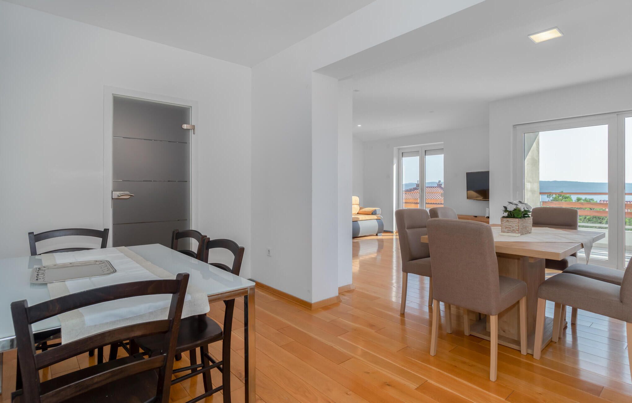 Uma sala de jantar moderna com uma mesa e cadeiras de madeira, um armário cinzento e vista para a cidade através de grandes janelas.