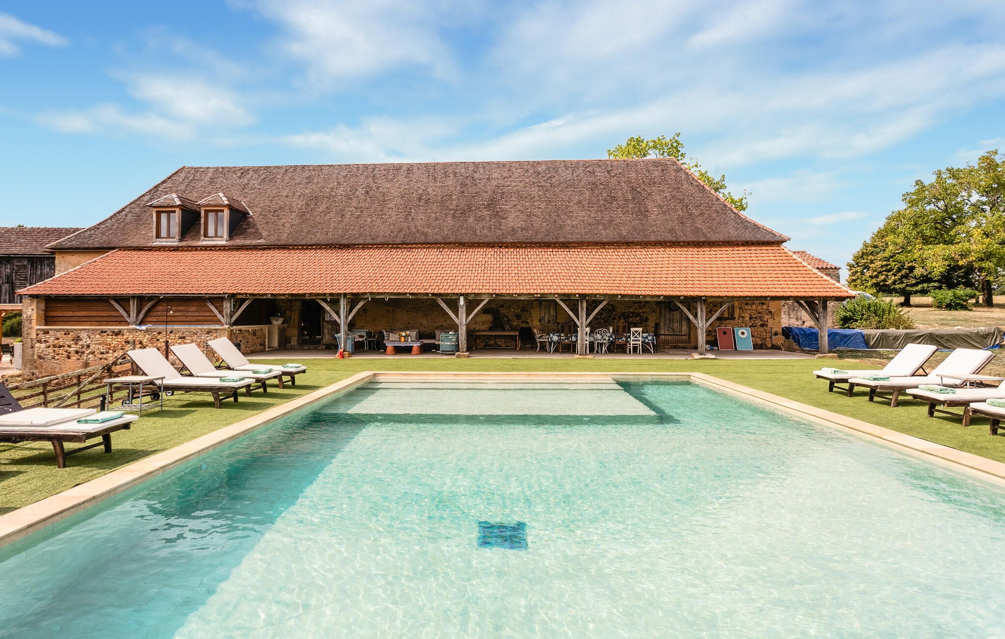 A pool area with lounge chairs, a covered patio, and a stone building with a tiled roof.