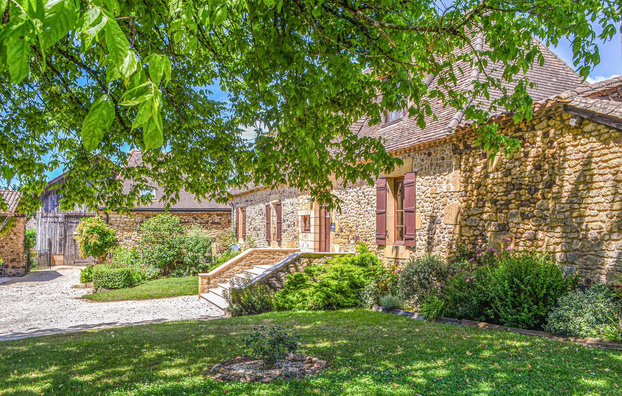 A stone building with a tiled roof, a stone staircase, and a well-maintained garden with various plants and a gravel path.