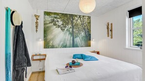 A bedroom with a large forest-themed painting, a bed with blue pillows, a bedside table with a lamp, and a window with a view of trees.