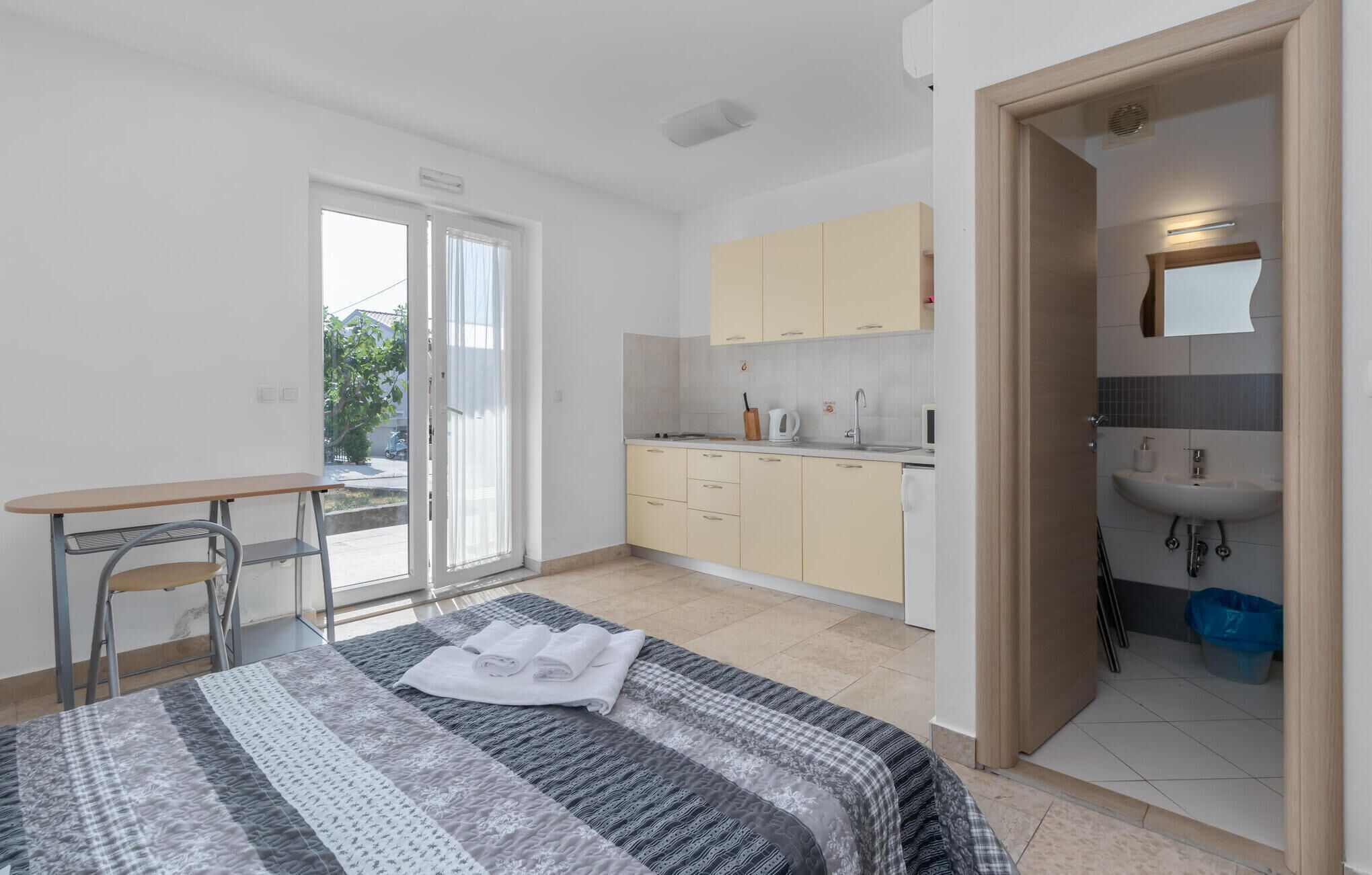 Een moderne, goed verlichte slaapkamer met een bed, een klein bureau en uitzicht door een schuifdeur op een keuken.