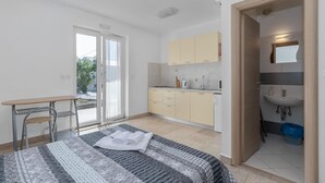 Une chambre à coucher moderne et bien éclairée, avec un lit, un petit bureau et une vue sur un espace cuisine accessible par une porte coulissante.
